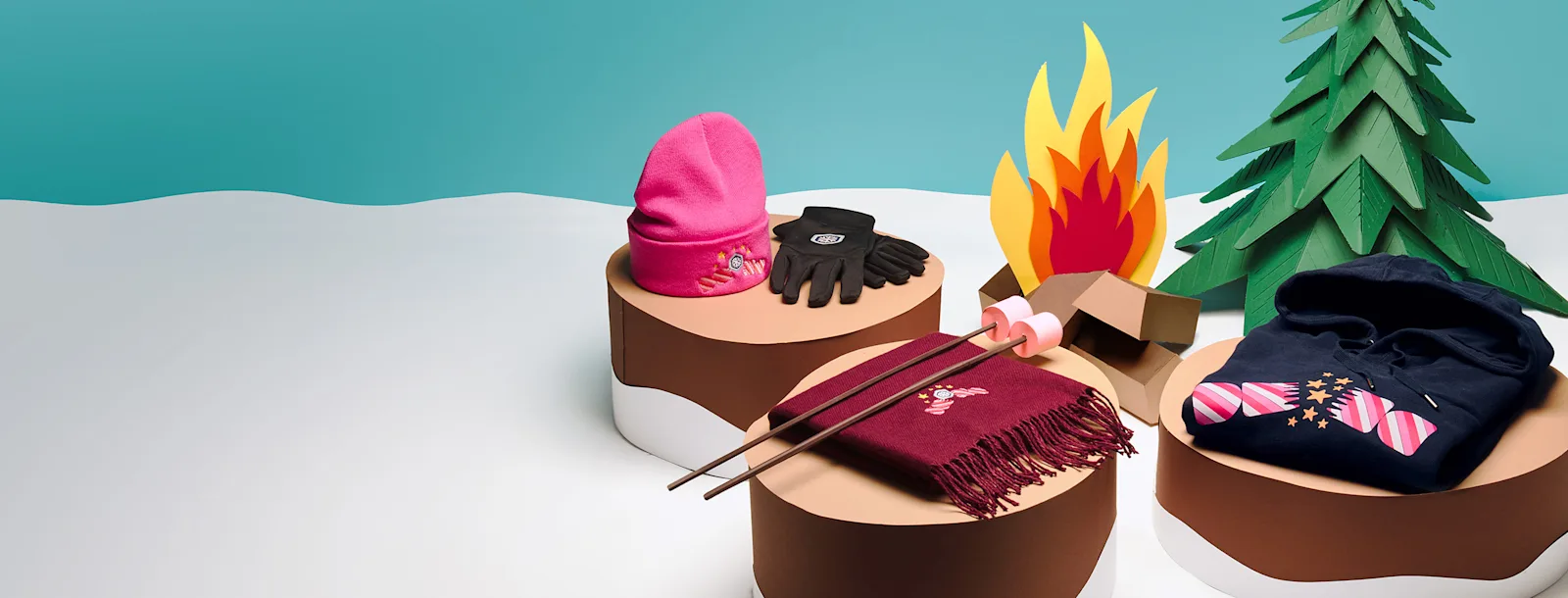 Haube, Handschuhe, Schal und Jacke mit Logo auf Baumstümpfen im Schnee neben einem Lagerfeuer aus Papier